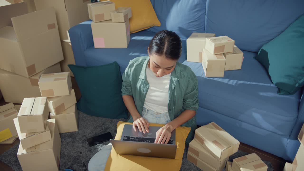 年轻的亚洲女售货员在网上商店使用笔记本电脑准备包裹发货给客户在家里。