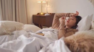 一名亚洲华裔妇女躺在床上，盖着毯子，患感冒和流感，用智能手机和朋友聊天，用数字体温计测量体温视频素材模板下载