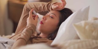 一名亚洲华裔妇女躺在床上，盖着毯子，患感冒和流感，用智能手机和朋友聊天，用数字体温计测量体温