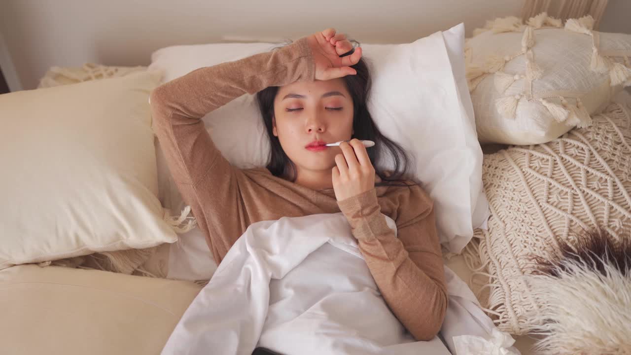 一名亚洲华裔妇女躺在床上，盖着毯子，患感冒和流感，用智能手机和朋友聊天，用数字体温计测量体温