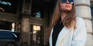 一个戴着眼镜，穿着白色外套的女孩走过城市的商业区。在豪华轿车和公寓的背景下