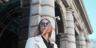 一个戴着眼镜、穿着白色外套的女孩站在市中心一家酒店的背景下，调整着自己的眼镜