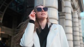 一个戴着眼镜、穿着白色外套的女孩站在市中心一家酒店的背景下，调整着自己的眼镜视频素材模板下载