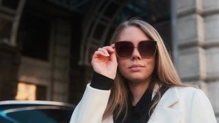 一个戴着眼镜、穿着白色外套的女孩站在市中心一家酒店的背景下，调整着自己的眼镜视频素材模板下载