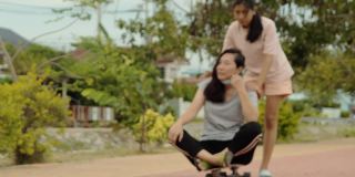 快乐的亚洲女孩推动她的母亲谁坐在滑板与乐趣一起户外，生活方式的概念。