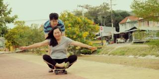 快乐的亚洲男孩推着他的母亲谁坐在滑板与乐趣一起户外，生活方式的概念。