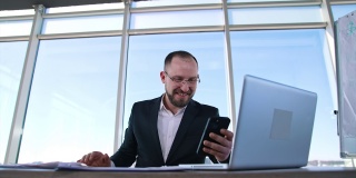 微笑的商人正在用他的手机打视频电话。戴眼镜、穿西装的上班族坐在桌子旁，用笔记本电脑打手机。