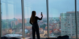长头发的女孩在屋里拿着电话。一名年轻女子在办公室窗口拍摄美丽的城市夜景。后面的观点。