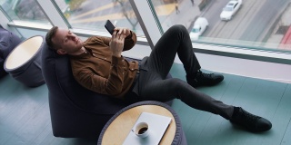 一个男人拿着电话，坐在柔软的椅子上休息。一个年轻人坐在窗边，有城市交通的背景，在用他的手机。小桌子上有一杯咖啡。前视图。