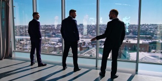 严肃的商人站在窗边。穿着西装的优雅企业家在摩天大楼里听他们的伙伴说话。后视图。
