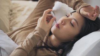 一名亚洲华裔妇女躺在床上，盖着毯子，患感冒和流感，用智能手机给朋友发短信，用数字温度计测量体温视频素材模板下载