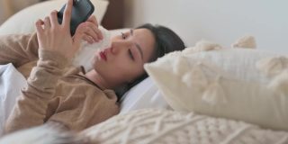 一名亚洲华裔妇女躺在床上，盖着毯子，患感冒和流感，用智能手机给朋友发短信，用数字温度计测量体温