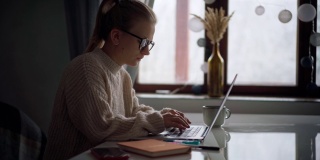 一个年轻的女孩戴着眼镜在笔记本电脑上打字，在笔记本上做笔记