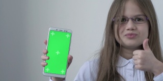 一名身穿白色校服的女学生用绿色屏幕模拟手机、手机、电话。绿屏智能手机的色度键设置为广告。教育、科技、小玩意和孩子