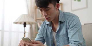 一名困惑的亚洲男子在家中客厅查看手机上令人震惊的通知信息时，手掌向上看着屏幕的特写镜头。