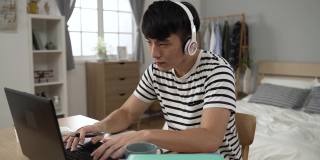 一名亚裔男生戴着耳机，全神贯注地在卧室里写笔记，在笔记本电脑上打字
