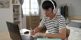 一名亚洲大学生戴着耳机，面带微笑，在家里通过电脑在线学习的同时，通过视频与老师交谈