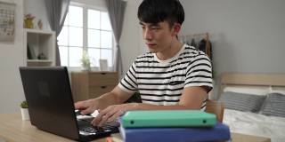 集中注意力的亚洲男学生一边在卧室里做作业，一边在家里学习，一边在电脑上搜索信息，做笔记。