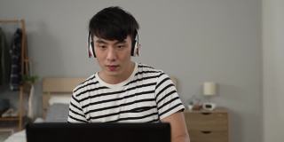 一个快乐的亚洲年轻人在家里的电脑上看在线音乐视频的时候，一边听着耳机里的歌曲，一边享受着前方镜头的慢动作。