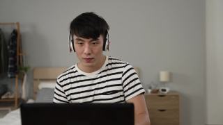 一个快乐的亚洲年轻人在家里的电脑上看在线音乐视频的时候，一边听着耳机里的歌曲，一边享受着前方镜头的慢动作。视频素材模板下载