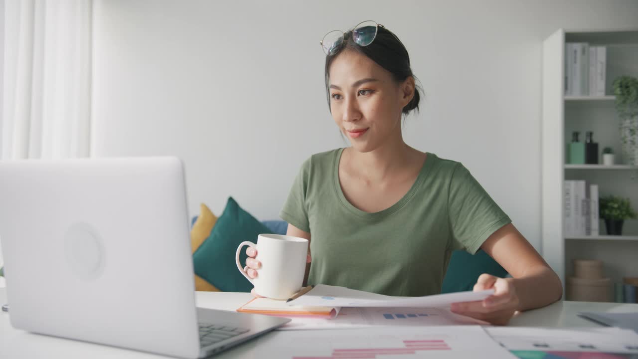 亚洲商务女性穿着休闲服装，在家里的客厅里用笔记本电脑进行视频会议，喝咖啡。