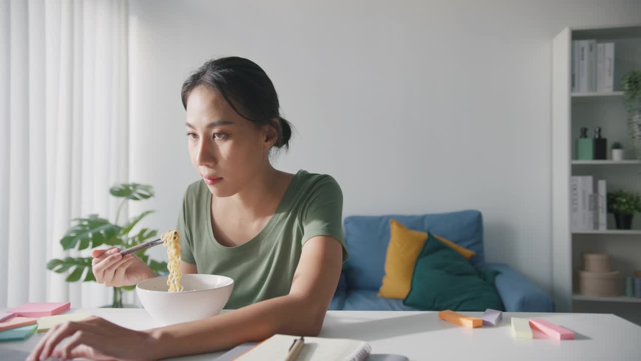 亚洲商务女性，穿着休闲，吃方便面，在家里客厅用笔记本电脑工作。