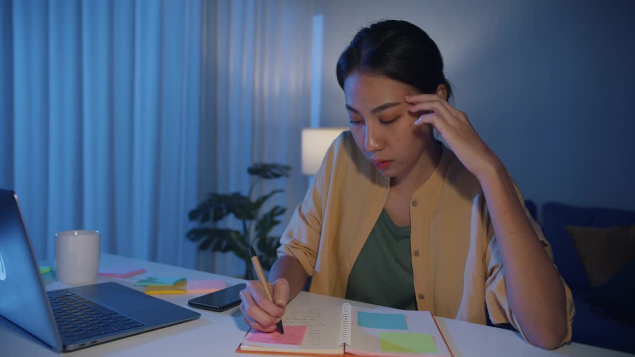 疲倦的年轻亚洲商业妇女在休闲工作笔记本电脑在客厅在家里。