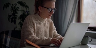 一个年轻的女孩戴着眼镜在笔记本电脑上打字，在笔记本上做笔记