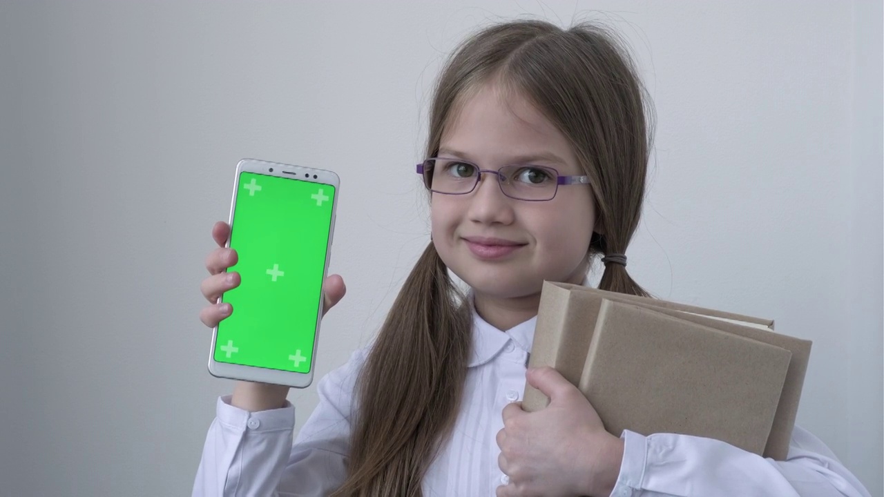 一名女学生，穿着白色的校服，戴着眼镜，拿着书，拿着绿屏手机。绿屏智能手机的色度键设置为广告。教育、科技、小玩意和孩子