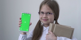 一名女学生，穿着白色的校服，戴着眼镜，拿着书，拿着绿屏手机。绿屏智能手机的色度键设置为广告。教育、科技、小玩意和孩子