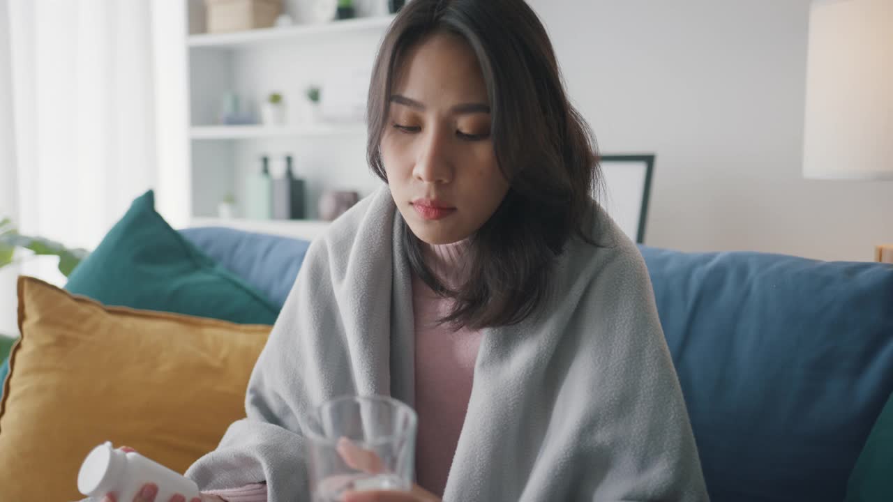 生病的亚洲妇女喝水喝处方药，在家里客厅的沙发上盖着毯子，忍受着头痛或抑郁。