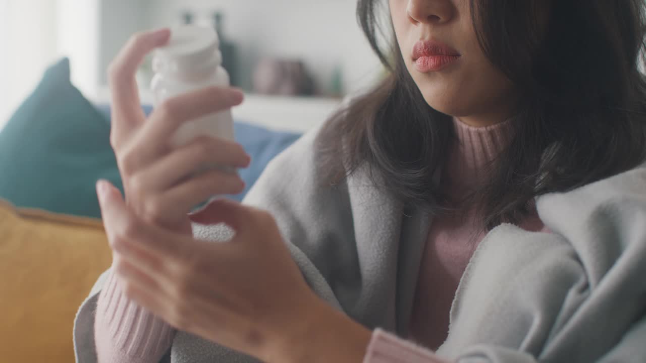 生病的亚洲妇女喝水喝处方药，在家里客厅的沙发上盖着毯子，忍受着头痛或抑郁。近距离射杀。