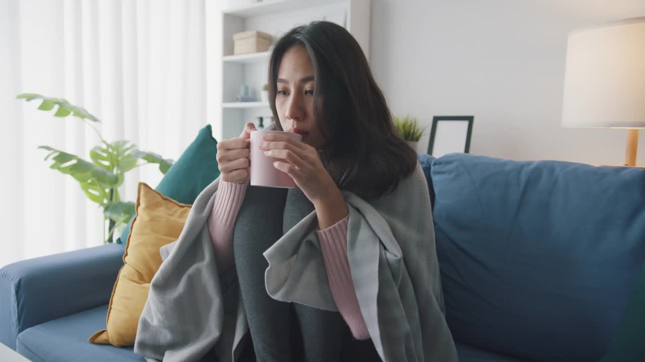 生病的亚洲妇女喝热水坐在沙发上的毯子在客厅在家里。