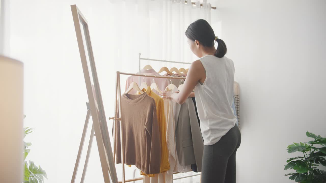 年轻的亚洲妇女在衣架上挑选衣服，在家里的客厅里穿衣照镜子。