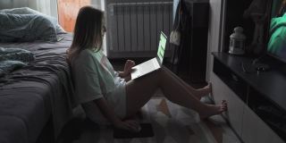 在一个多云的清晨，一名年轻女子正坐在电视机前的地毯上用笔记本电脑工作。她仰靠在摊开的沙发上。在家工作流行病商务距离