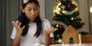 亚洲女孩舔手指上的糖，在晚上在家做圣诞饼干和姜饼屋，节日的生活方式概念。