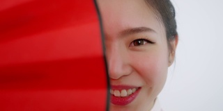 中国/亚洲美女春节脸的特写，红色扇子遮住了她的右半边脸，微笑着看着镜头