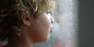 一个小男孩在寒冷的雨天站在窗边想出去