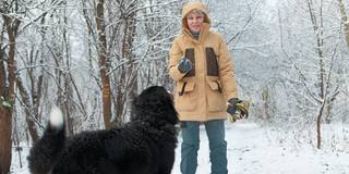 一个女人穿着一件黄色的冬季夹克，在雪原森林里遛着她的伯尔尼山狗。这只狗正从摄像机前跑向它的主人。缓慢的运动。