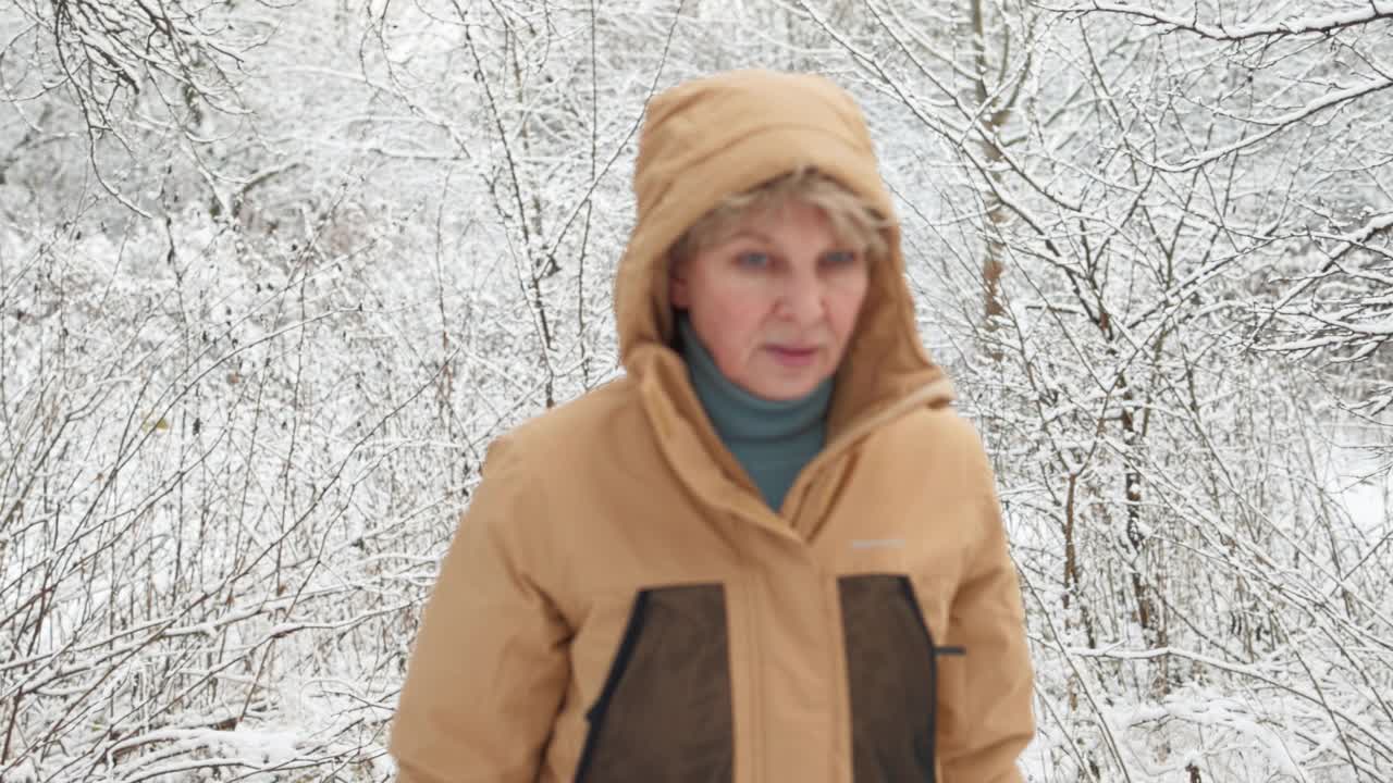 穿着黄色冬季夹克，戴着兜帽的成熟女性正在雪地里散步。选择性聚焦，剪辑开始和结束与离焦。