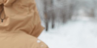 从离焦到离焦。成熟的女人穿着黄色的冬季夹克，头戴兜帽，走在雪地里的公园里。