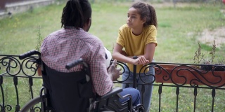 青春期的非洲裔美国女孩用慢动作和坐在轮椅上的残疾人交谈，他们正在踢足球。十几岁的女儿和父亲在后院户外休息的肖像。