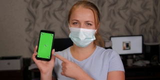 一名戴着医用口罩的妇女在家中手持一部绿色屏幕的手机。