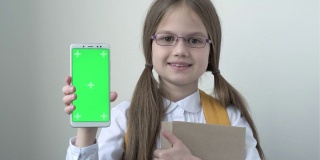 一名女学生，背书包，穿着白色的校衫，戴着眼镜拿着书，拿着绿色屏幕的手机。绿屏智能手机的色度键设置为广告。教育、技术和孩子