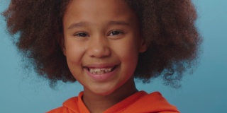 近距离的微笑非洲裔美国女孩穿着橙色的衣服看着镜头在蓝色的背景。7岁的快乐孩子。
