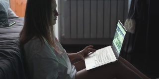 在一个多云的清晨，一名年轻女子正坐在电视机前的地毯上用笔记本电脑工作。她仰靠在摊开的沙发上。在家工作流行病商务距离