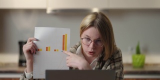 一位戴着眼镜的年轻女子坐在家里的厨房里，对着笔记本电脑的摄像头展示图表。家庭办公室远程工作。视频电话和会议与图形和表格。解释图