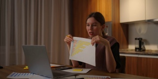 远程工作一位年轻女性坐在家中厨房里进行在线报告时，正在用笔记本电脑摄像头展示一幅图表。视频电话和会议演示文件和时间表