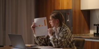 家庭办公室一位戴眼镜的年轻女子坐在厨房里，对着笔记本电脑的摄像头展示图表，进行在线报告。视频电话和会议演示文件