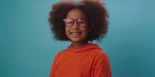 微笑的非洲裔美国女孩，戴着眼镜，穿着橙色的衣服，看着镜头在蓝色的背景。7岁戴眼镜的快乐小孩。
